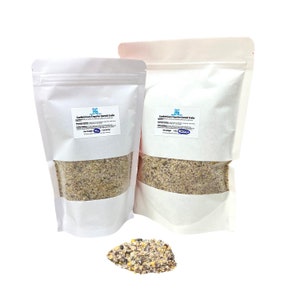 May include: Two white bags of Confident Care Treat for Hermit Crabs. The bags are filled with a mixture of brown, white, and green food pellets. A small pile of the food pellets is in front of the bags.