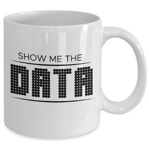 Op de afbeelding: Witte keramische koffiemok met een zwart ontwerp met de tekst "Toon me de data" met het woord "data" in een pixellettertype.