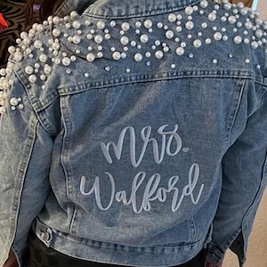 May include: A light blue denim jacket with white pearl embellishments on the shoulders and the text "Mrs. Walford" embroidered on the back.