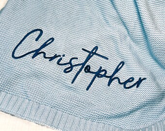 May include: Light blue knitted baby blanket with the name "Christopher" embroidered in navy blue thread. The blanket is folded and a brown stuffed animal is partially visible in the background.