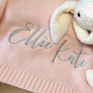 May include: A pink knitted baby blanket with the name "Ellie Kate" embroidered in silver thread. A white plush bunny is lying on top of the blanket.