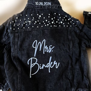 May include: Black denim jacket with pearl embellishments on the shoulders and the text "Mrs. Binder" embroidered on the back in white. The date "10.26.2024" is written on the collar.