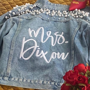 May include: A light blue denim jacket with white pearl embellishments on the shoulders and the words "Mrs. Dixon" embroidered on the back.
