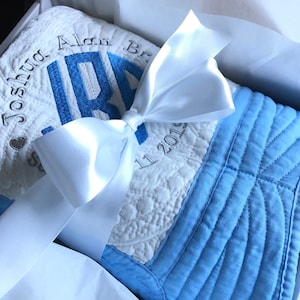 May include: A white and blue quilted baby blanket with a white satin ribbon tied in a bow. The blanket is embroidered with the name "Joshua Alan" and the date "Sept 11, 2015".
