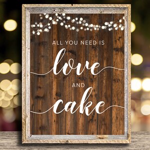 May include: A rustic wooden sign with a white frame and string lights. The sign says "All you need is love and cake".