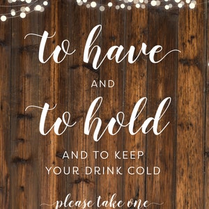 May include: A rustic wooden sign with white text that reads "to have and to hold and to keep your drink cold please take one". The sign is decorated with a string of white lights.