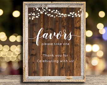 May include: A rustic wooden sign with a white frame and string lights. The sign says "favors please take one" and "Thank you for celebrating with us!"