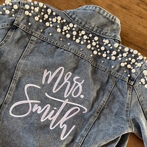 May include: A blue denim jacket with white pearl embellishments around the collar and shoulders. The back of the jacket has the words "Mrs. Smith" embroidered in white.