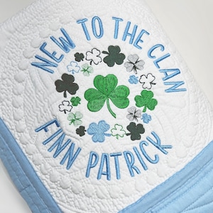 May include: A white quilted blanket with a light blue border featuring embroidered text that reads NEW TO THE CLAN FINN PATRICK in light blue. The center design includes a circular arrangement of various green and blue shamrock icons.