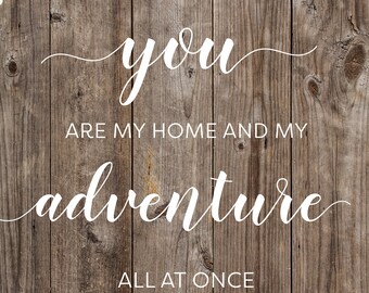 May include: A rustic wooden sign with the words "you are my home and my adventure all at once" in white script. The background is made of vertical wooden planks, and there are string lights at the top.