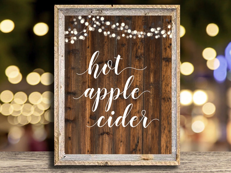 Puede incluir: Un letrero de madera r&uacute;stico con texto blanco que dice "hot apple cider". El letrero est&aacute; decorado con una cadena de luces blancas.