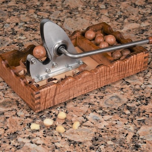 May include: A wooden tray with a metal nutcracker and macadamia nuts. The nutcracker has a long handle and a metal blade. The tray is made of wood with a natural finish. The macadamia nuts are in the tray and on the counter.