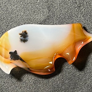 Può includere: Una pietra di agata bianca, arancione e marrone con motivi dendritici neri. La pietra è scolpita in una forma libera con una superficie liscia e lucida.