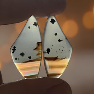 Puede incluir: Dos gemas translúcidas en forma de lágrima se sostienen entre los dedos. Las piedras tienen una base gris claro con inclusiones negras y bandas de color naranja y amarillo. El fondo está borroso con tonos cálidos y dorados.