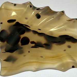 Può includere: Una pietra di agata beige e marrone con una forma ondulata organica e inclusiones nere e marroni. La pietra è lucidata e ha una superficie liscia.