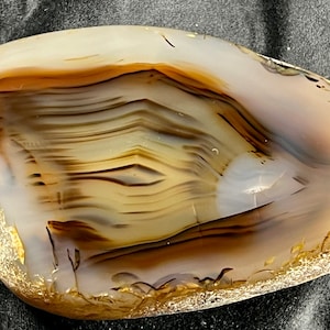 Può includere: Una pietra di agata lucidata con una forma ovale e liscia. La pietra presenta bande marroni, beige e bianche, creando un motivo a strati. I bordi hanno un aspetto ruvido e strutturato. Questa pietra naturale è un oggetto decorativo.