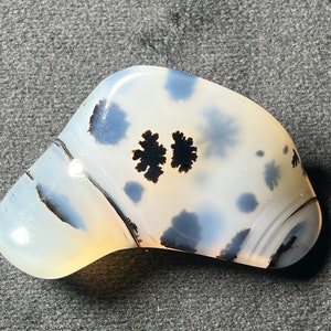 Può includere: Una pietra di agata bianca con motivi dendritici neri. La pietra è liscia e lucidata, con una forma leggermente curva.