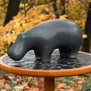 May include: A black ceramic hippopotamus sculpture on a small, round wooden table with a mosaic tile top. The table has a light brown wooden rim and a single, slender leg. The background features a blurred view of autumn foliage.