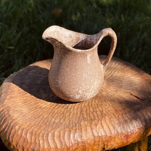 May include: A small, brown ceramic pitcher with a handle and a wide spout. The pitcher has a speckled texture and sits on a carved wooden stool. The background is blurred green grass, suggesting an outdoor setting. The pitcher is likely for decorative use.
