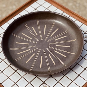 May include: A brown ceramic plate with a sunburst design in cream-colored lines. The plate is round and has a slightly raised edge. The plate is sitting on a white tiled surface with a wooden border.