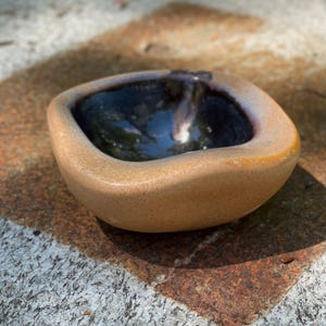 May include: A small, handmade ceramic bowl with a square shape and rounded edges. The exterior is a speckled tan color, while the interior is a glossy, dark blue. The bowl sits on a textured, stone surface.