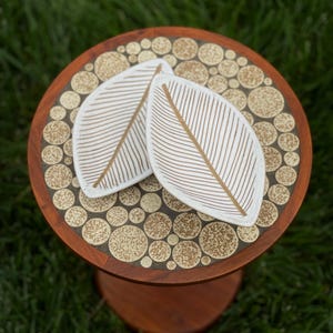 May include: A small, round wooden table with a decorative top featuring a mosaic of beige circles. Two white leaf-shaped coasters with gold-colored vein details rest on the table. The table has a single, rounded leg.