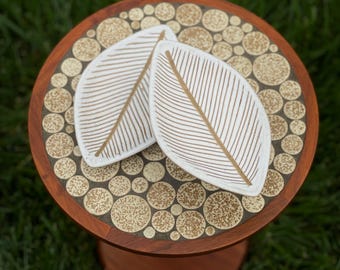 May include: A small, round wooden table with a decorative top featuring a mosaic of beige circles. Two white leaf-shaped coasters with gold-colored vein details rest on the table. The table has a single, rounded leg.