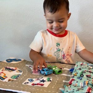 May include: A child plays with toy cars on a road play mat. The mat features a road design with various vehicles and buildings. The child is wearing a white t-shirt with a red trim and a graphic print.