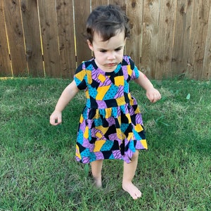 May include: A young child wearing a colorful patchwork dress with a black, yellow, purple, and blue pattern. The dress has a short sleeve and a ruffled skirt. The child is standing on a grassy lawn.