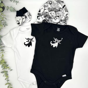 May include: A set of baby clothing including one white bodysuit and one black bodysuit, both featuring a white graphic of a ghost dog. The set includes a pair of matching black and white patterned baby booties and a matching knotted baby hat.