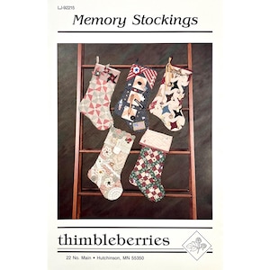 May include: Five quilted Christmas stockings labeled Memory Stockings by thimbleberries. The stockings feature various fabric patterns including red and white pinwheels, navy blue with stars, beige with brown stars, cream lace, and a green and red grid.