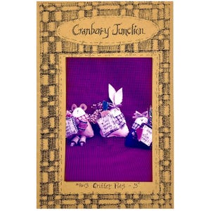 May include: A framed image featuring three fabric critter pins. The pins include a mouse and two bunnies, each holding a small sign with text. The frame has a plaid pattern and the text "Cranberry Junction".