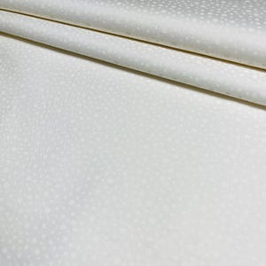 May include: Off-white fabric with a subtle, all-over polka dot pattern. The fabric is folded, showing the texture and drape of the material. The dots vary in size and are evenly spaced across the surface.