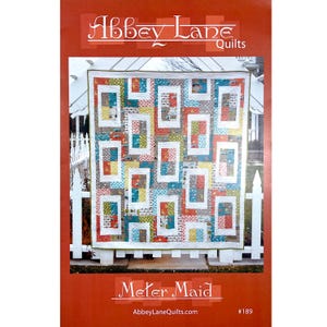 May include: A quilt with a geometric design in shades of red, blue, yellow, and white. The quilt is displayed on a white picket fence. The text "Abbey Lane Quilts" and "Meter Maid" are visible.