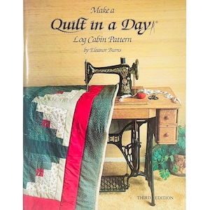 May include: A vintage sewing machine sits atop a wooden table, with a partially completed quilt draped over the edge. The quilt features a log cabin pattern with red, green, and white squares. The book cover reads "Make a Quilt in a Day" by Eleanor Burns.