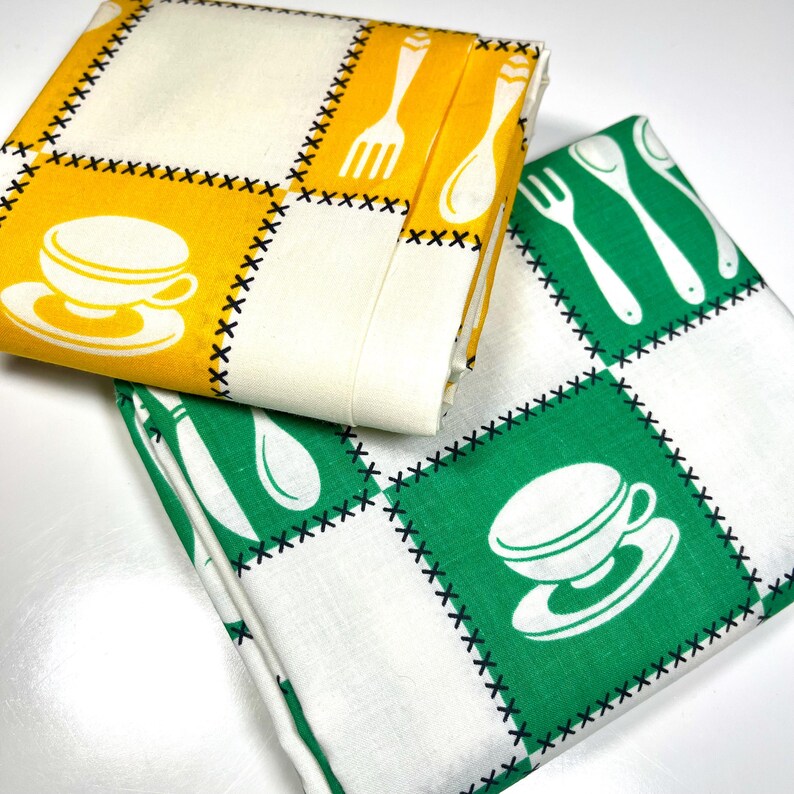 May include: Two folded kitchen towels with a retro design. One towel features yellow squares with white teacups, forks, and spoons. The other towel has green squares with white teacups and utensils. Both have black stitched borders.