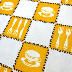 May include: A close-up of a white and yellow checkered tablecloth. The yellow squares feature white illustrations of coffee cups, forks, and knives. The squares are outlined with black stitching, creating a vintage, retro aesthetic.