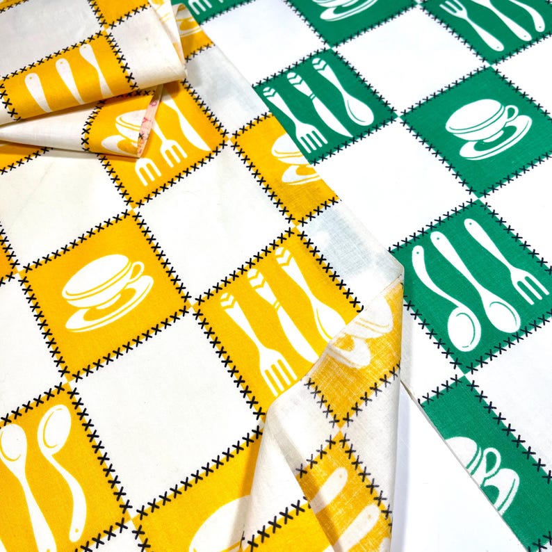 May include: A vintage kitchen tablecloth with a repeating pattern of white utensils and teacups on yellow and green squares. The squares are outlined with black stitching. The fabric is folded over in the image.