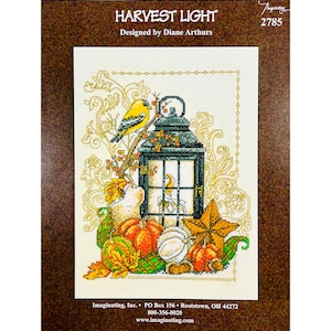 May include: Cross stitch pattern featuring a lantern with a yellow bird perched on a branch, surrounded by pumpkins, gourds, and fall leaves. The pattern is titled "Harvest Light" and designed by Diane Arthurs.