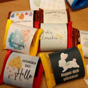 Könnte beinhalten: Mehrere Schokoriegel, verpackt in buntem Papier mit Ostermotiven. Die Verpackungen zeigen Bilder von Hasen, Küken und Eiern sowie Sätze wie "Ei-mazing!" und "Hallo". Die Riegel sind rot und gelb.