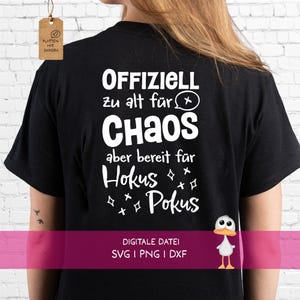 Op de afbeelding: Een zwart t-shirt met witte tekst op de achterkant: Offiziell zu alt für Chaos aber bereit für Hokus Pokus. Het ontwerp bevat een kleine tekstballon met een x en verschillende stervormige symbolen. Er is een klein bruin label bevestigd.