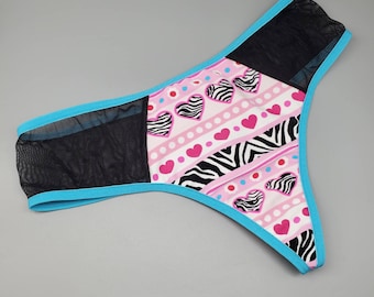May include: Pink and black patterned thong underwear with a turquoise trim and a mesh panel. The pattern features hearts, zebra stripes, and polka dots.