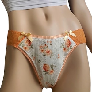 Puede incluir: Ropa interior con estampado floral blanco y naranja, con ribete de cuadros vichy. La ropa interior presenta detalles de encaje naranja y pequeños lazos amarillos. El estampado floral incluye rosas y hojas verdes.