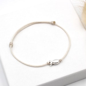 Op de afbeelding: Een wit koord armband met een zilveren vis bedel. De armband ligt op een wit oppervlak met een gedroogde grassoort op de achtergrond.