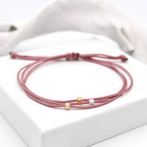 Op de afbeelding: Een roze gevlochten koord armband met drie kleine kralen in goud, zilver en rosé goud.