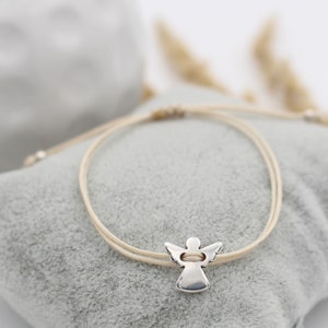 Peut inclure: Un bracelet en cordon beige avec un pendentif ange en argent. Le bracelet est posé sur une surface en tissu gris clair.
