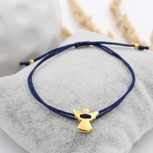 blaues Makramee Armband mit goldfarbenem Schutzengel auf grauem Kissen gelegen