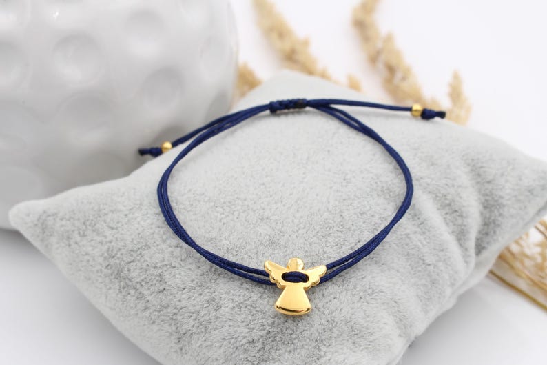 blaues Makramee Armband mit goldfarbenem Schutzengel auf grauem Kissen gelegen