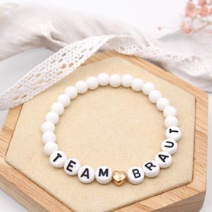 Könnte beinhalten: Ein kreisförmiges Perlenarmband mit matten, weißen, runden Perlen. Das Armband enthält schwarze Buchstabenperlen, die den Text TEAM BRAUT bilden, getrennt durch eine kleine, metallisch goldfarbene herzförmige Perle in der unteren Mitte.