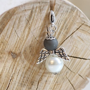 Könnte beinhalten: Ein silberner Engelsanhänger mit grauen und weißen Perlen. Der Engel hat Flügel und eine perlmuttartige Perle in der Mitte.